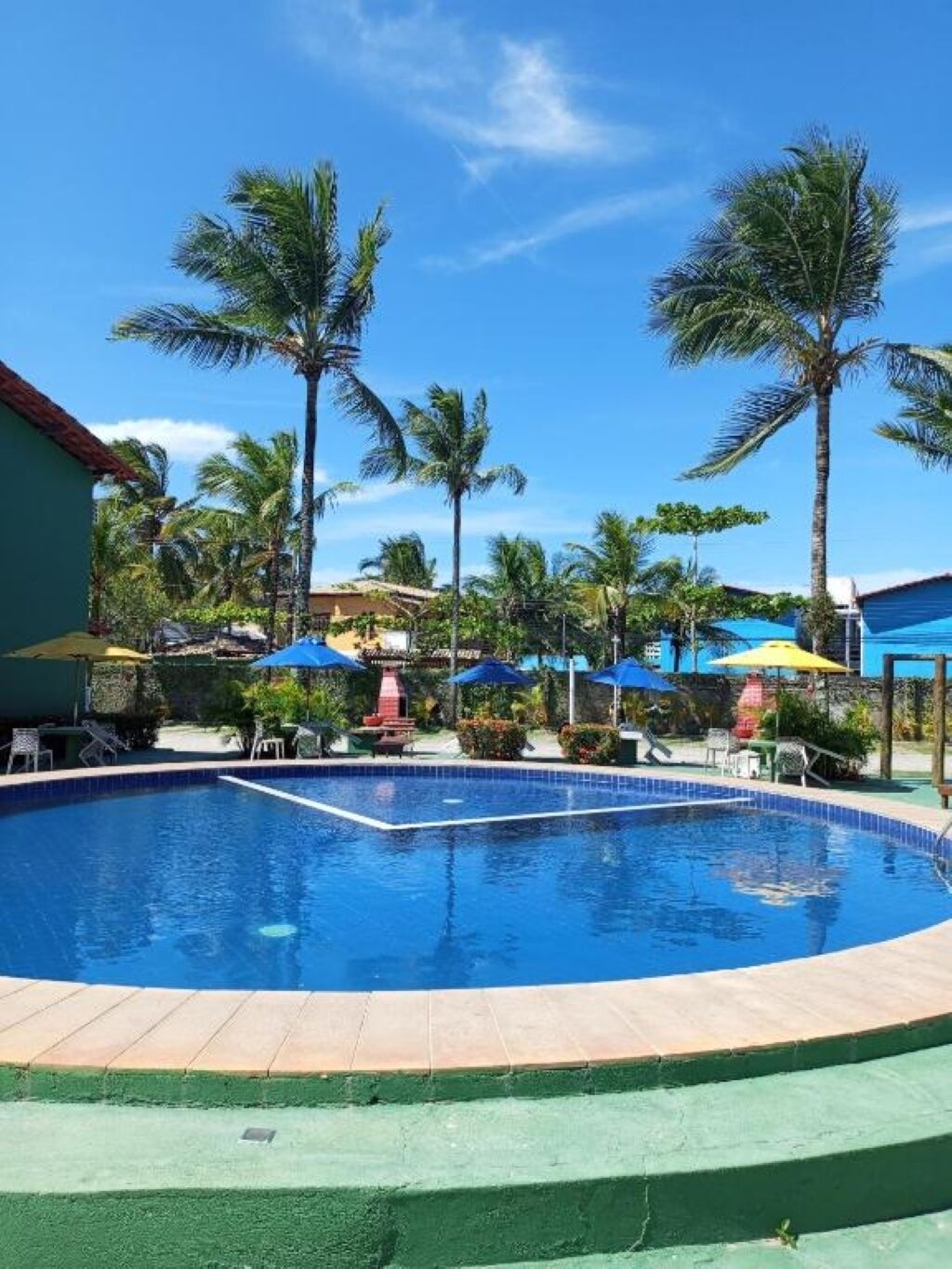 Ein Swimmingpool mit fliesenförmiger Kante, umgeben von Palmen und Sonnenschirmen, was auf eine Resorteinrichtung hindeutet.