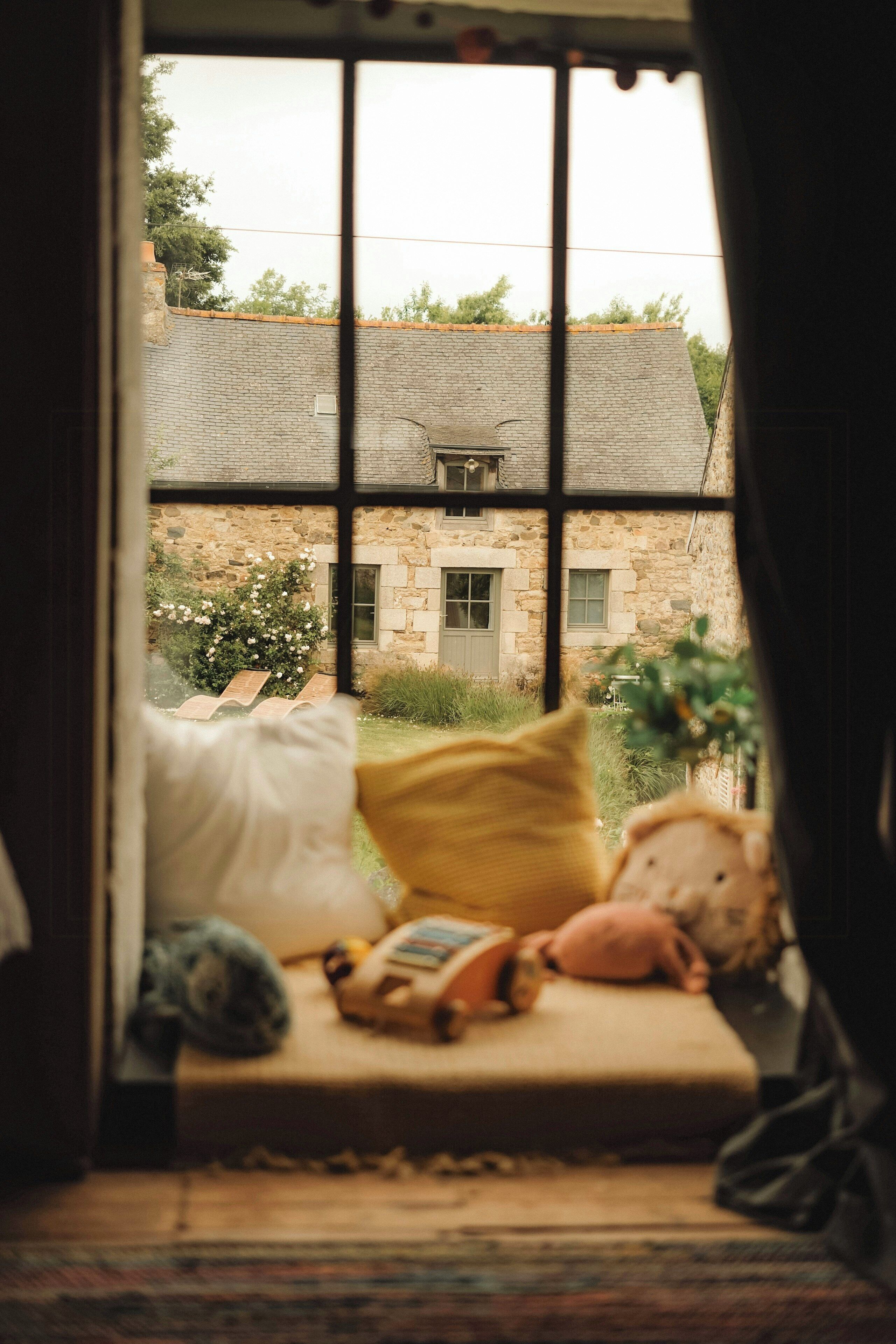 A room with a large window, a bed with pillows, and a view of a stone building outside.