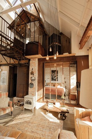 Ein Loft-Zimmer mit hoher Decke, freiliegenden Balken und einer Schlafgalerie, die über einer Holztreppe erreichbar ist.