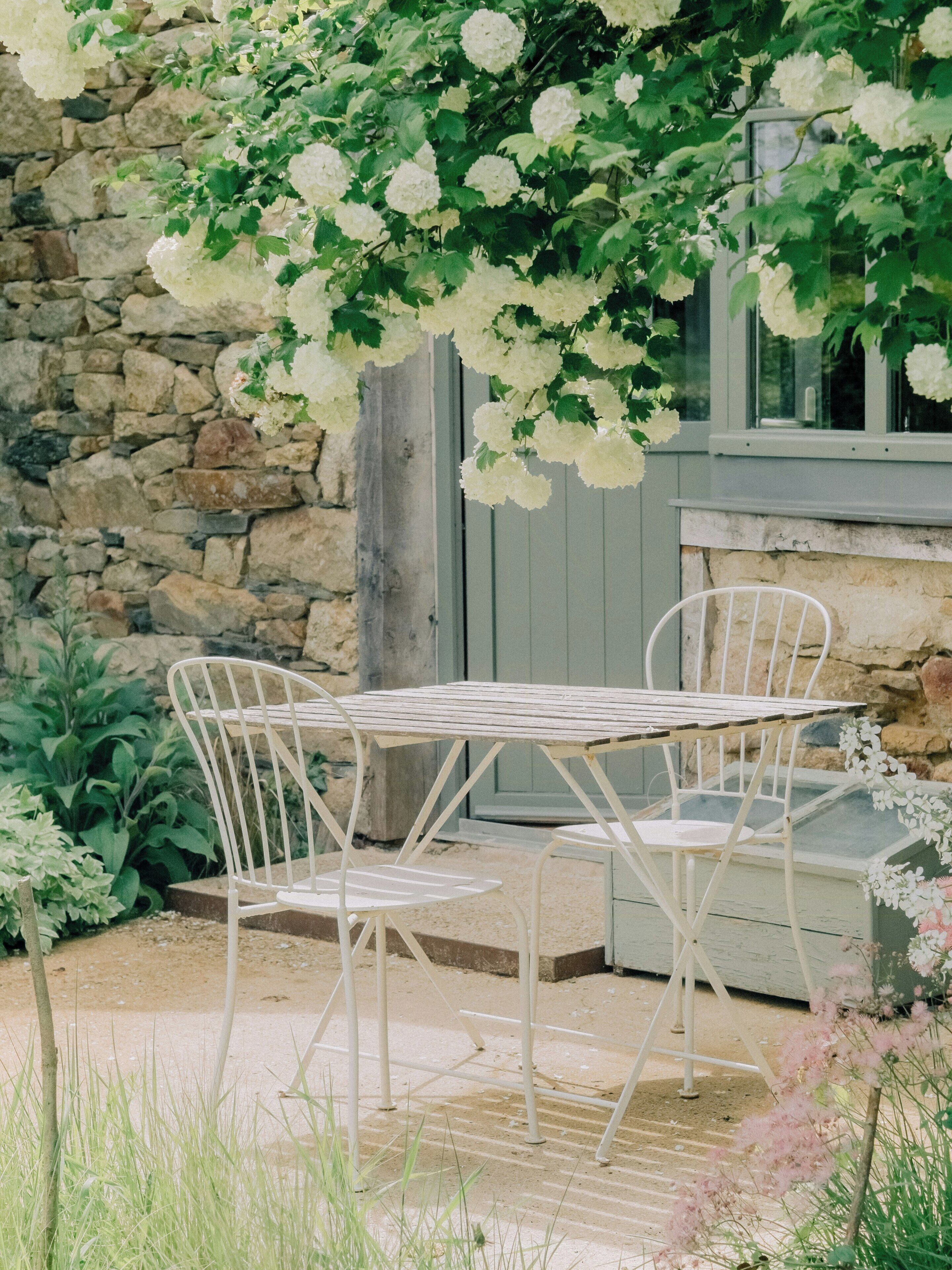 Een stenen muur, een witte metalen tafel met stoelen en witte bloemen.