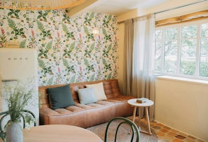Ein gemütliches Zimmer mit einer Couch, einem kleinen Tisch, einem Fenster mit Vorhängen und einem gemusterten Wandteppich.