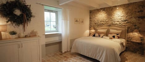 A bedroom with a stone wall, a bed with white bedding, a wooden headboard, a window with white curtains, and a white cabinet with a wreath.