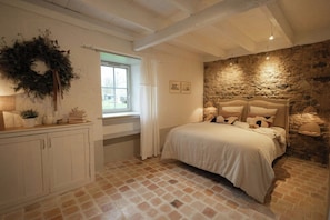 Ein Schlafzimmer mit Steinwand, einem Bett mit weißer Bettwäsche, einem Holz-Kopfteil, einem Fenster mit weißen Vorhängen und einem weißen Schrank mit einem Kranz.