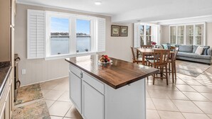 A kitchen with a central island, dining area with wooden chairs, a sofa, and large windows with shutters.