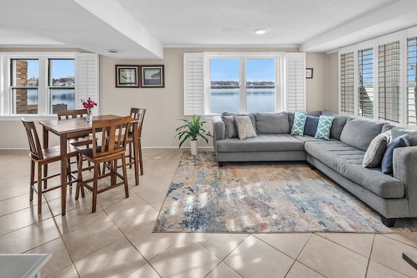 A living room with a dining table, gray sofa, and large windows overlooking a body of water.