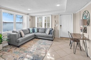 A corner living room with a gray sectional sofa, a small dining table with chairs, a patterned rug, and a view of a body of water through large windows.