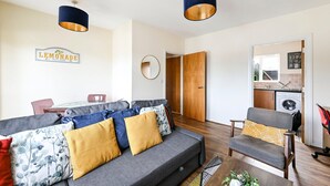 Een woonkamer met een bank, fauteuil en salontafel, een keukenhoek met een wasmachine, en een deur naar een badkamer.