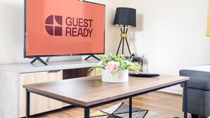 Een moderne woonkamer met een flatscreen televisie waarop 'GUEST READY' staat, een bijzettafeltje met een potplant, een vloerlamp en een bank.