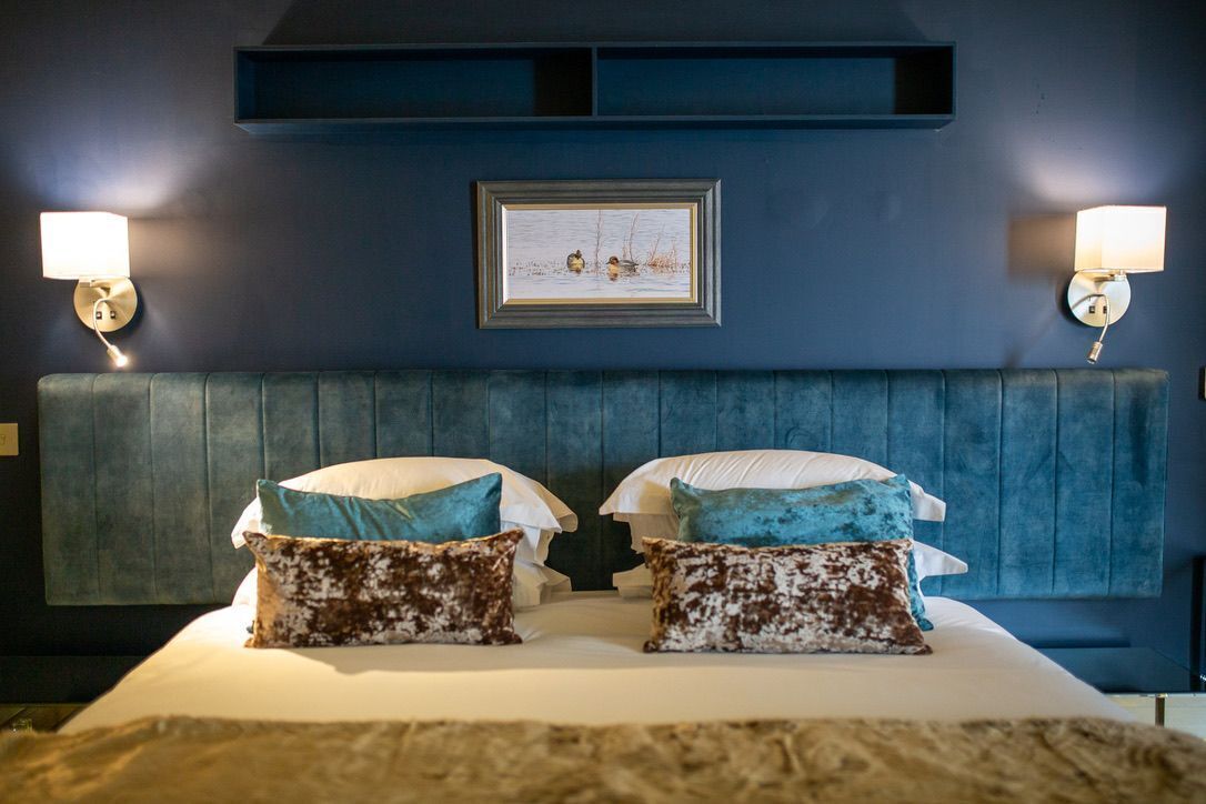 Un dormitorio con cabecera azul, dos lámparas de noche, un cuadro enmarcado y dos almohadas.