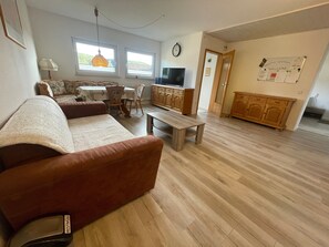 A living room with a sofa, a dining table with chairs, a TV, wooden cabinets, and a clock on the wall. - Apartment approx. 53 m² with Terrace (Niedereschach)