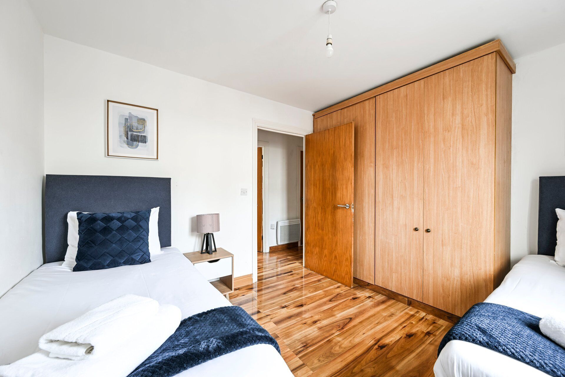 A hotel room with two beds, a large wardrobe, a framed picture on the wall, and a bedside table with a lamp.