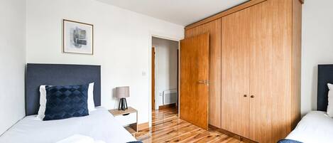 A hotel room with two beds, a large wardrobe, a framed picture on the wall, and a bedside table with a lamp.