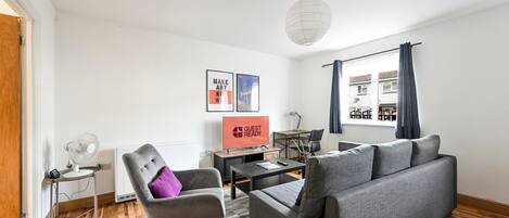 A modern living room with a grey sofa, a grey armchair, a small table, a TV, and a window with curtains.
