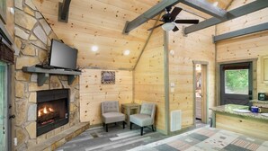 Interior de una cabaña de troncos con chimenea de piedra, televisor montado en una pared de piedra, dos sillones tapizados, ventilador de techo y una zona de cocina.
