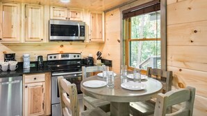 A wooden cabin interior with a kitchen featuring a microwave, stove, and cabinets. There is a dining table with chairs and a window with a view of greenery. - Lakeside Rendezvous (Gatlinburg)