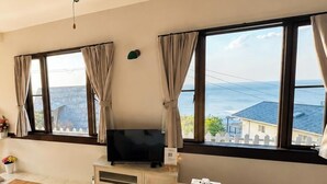 A living room with a TV, a sofa, a wooden table, and large windows with curtains. - Peaceful Ocean View Villa with Free Parking & Full Kitchen (Miyazaki)