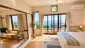 A hotel room with a large bed, a desk, a chair, a potted plant, and a view of the sea. - Peaceful Ocean View Villa with Free Parking & Full Kitchen (Miyazaki)