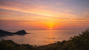 A sunset over a calm sea with a distant landmass and vegetation in the foreground. - Peaceful Ocean View Villa with Free Parking & Full Kitchen (Miyazaki)