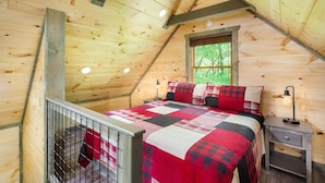 Une chambre dans une cabane en bois, avec un plafond en pente, un lit recouvert d’une couverture à carreaux rouges et noirs, une fenêtre donnant sur la verdure, et une petite table de chevet avec une lampe.