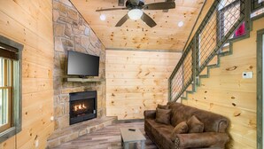 L’intérieur d’une cabane en rondins, avec une cheminée en pierre, un téléviseur, un canapé en cuir marron, un plancher en bois et un ventilateur de plafond.