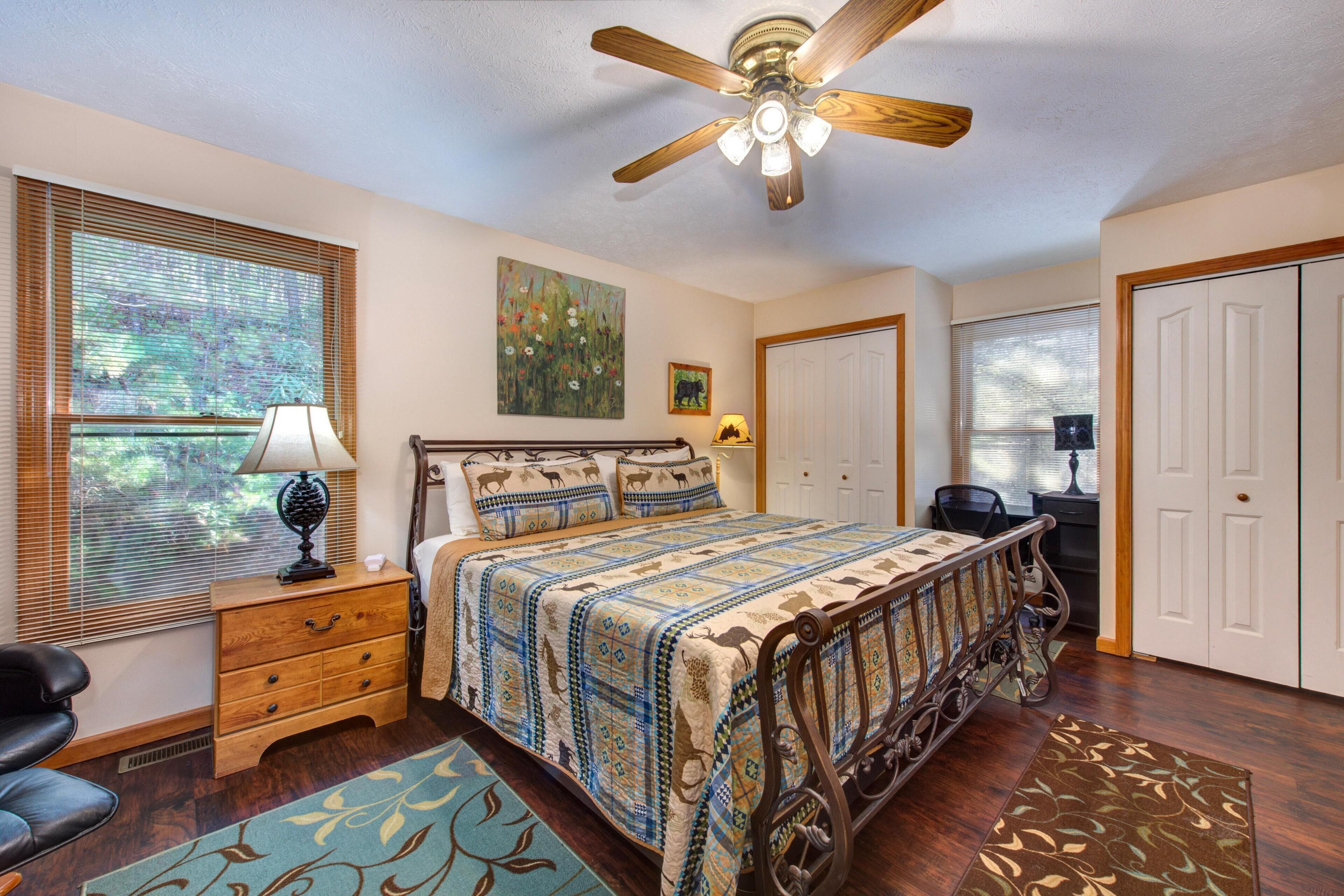 A bedroom with a bed, a nightstand, a lamp, a ceiling fan, a window with blinds, a door, and a chair.