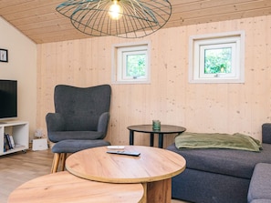 A living room with a grey sofa, a round wooden coffee table, a grey armchair, a TV on a white stand, and two windows with greenery visible outside. - 5 Star Holiday Home in Hovborg (Hovborg)