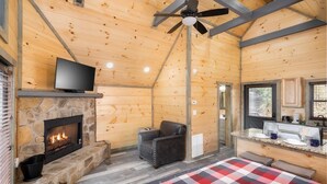 A log cabin interior with a stone fireplace, a television, a ceiling fan, a kitchen area, and a checkered rug. - Solitude Studio (Gatlinburg)