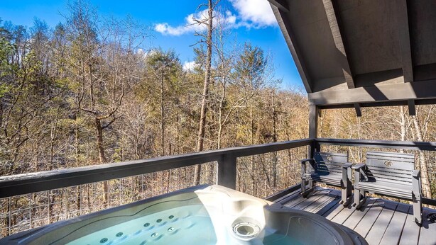A hot tub on a wooden deck with benches, surrounded by trees. - Solitude Studio (Gatlinburg)