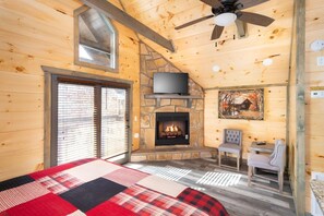 A log cabin room with a stone fireplace, a ceiling fan, a television, and a plaid bedspread. - Still Waters Studio (Gatlinburg)