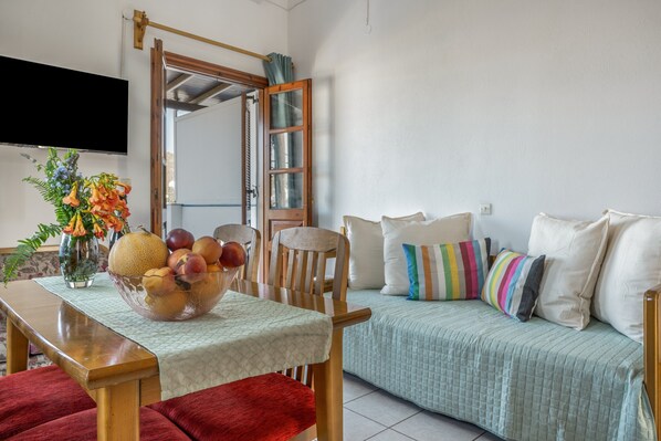 A room with a dining table, chairs, a sofa with colorful pillows, a fruit bowl, and a television on the wall. - Anastasia's 1 Apartment with Private Balcony and Sea View (Skala)