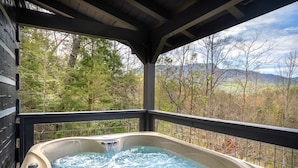 Un jacuzzi con caída de agua, rodeado por una terraza de madera y una baranda de metal, con vistas a un paisaje boscoso y montañas a lo lejos.