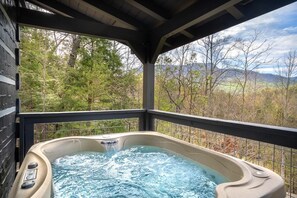 A hot tub with a waterfall feature, surrounded by a wooden deck and a metal railing, overlooking a forested landscape with mountains in the distance.