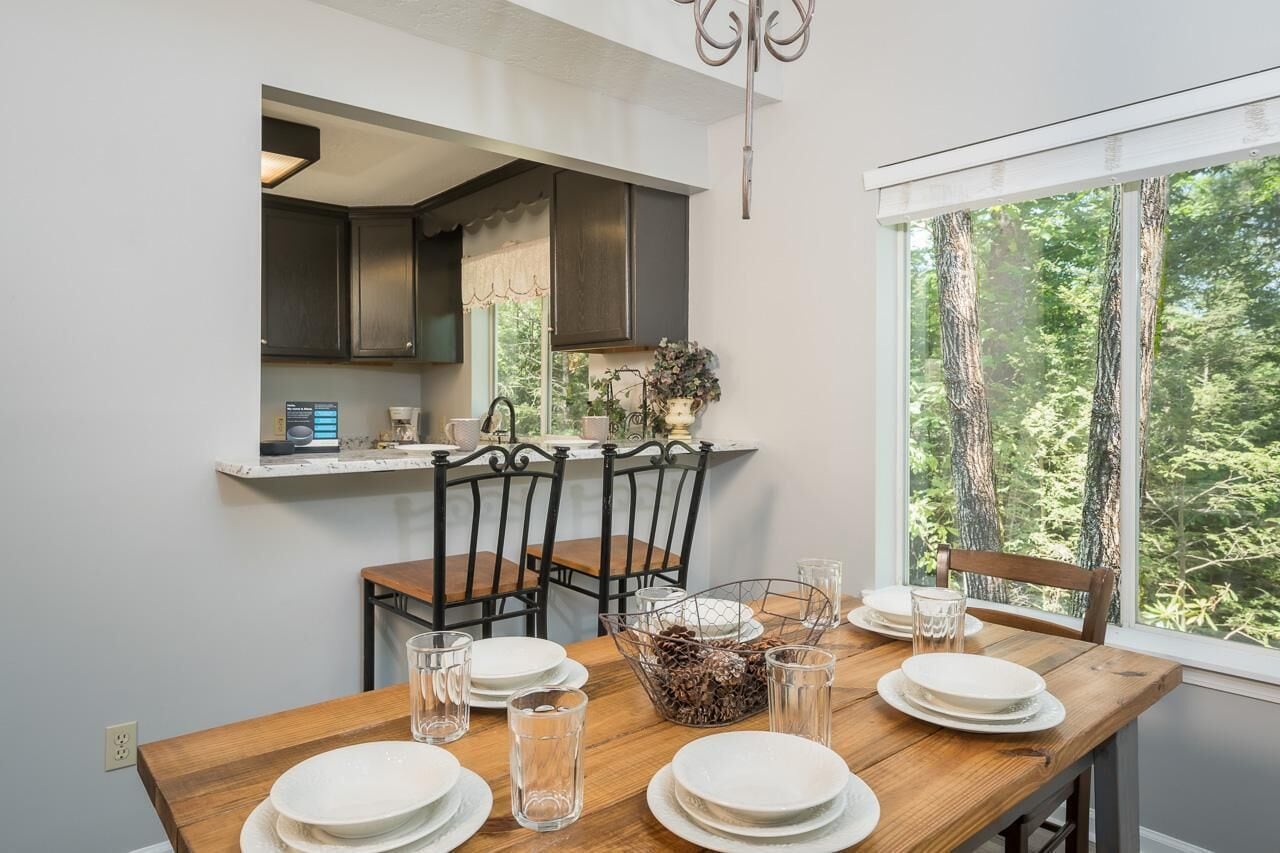 Un comedor con una mesa de madera dispuesta para cuatro personas, cuatro sillas, una ventana con vistas a un bosque y una cocina visible a través de una puerta abierta.