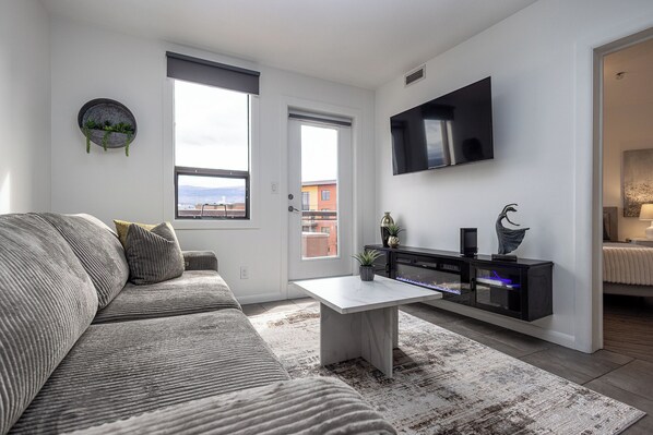 A modern living room with a grey sofa, a white coffee table, a flat-screen TV mounted on the wall, and a small plant on the side table. - Breathe taking resort in the heart of the Lower Mission. (Kelowna)