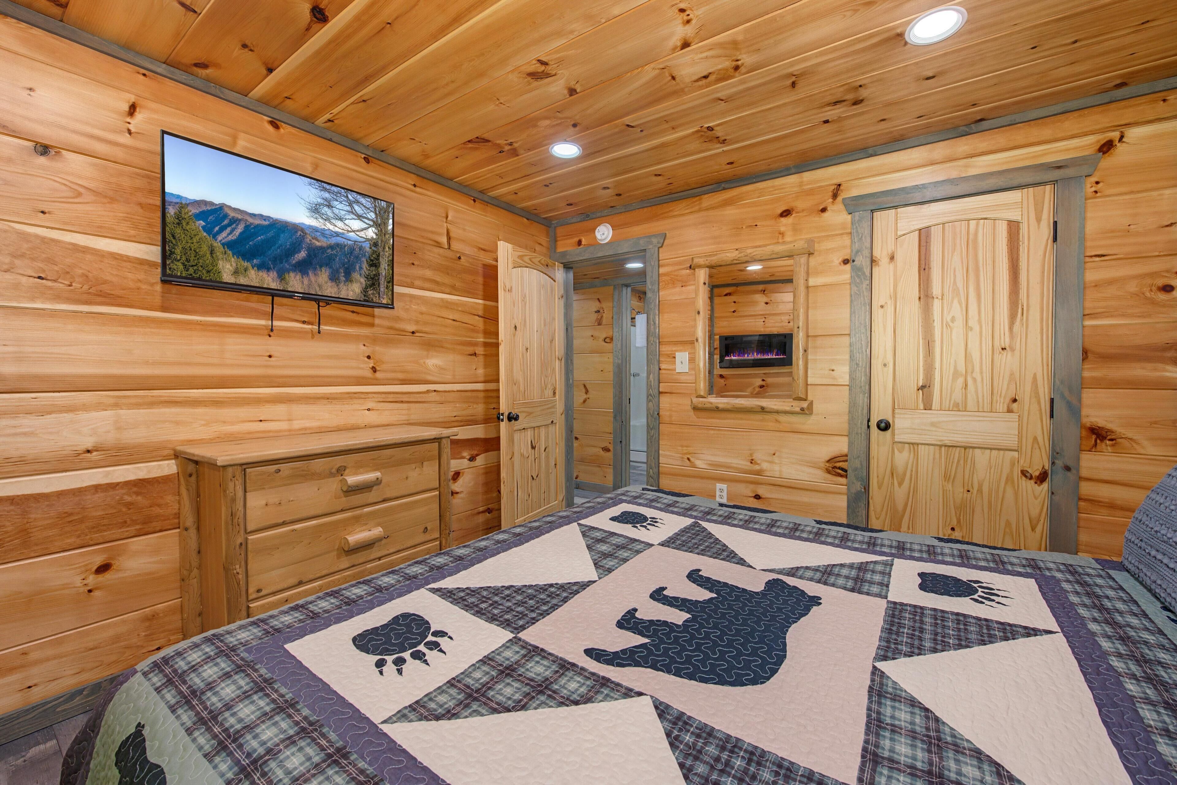 A wooden cabin interior with a bed, a television, a fireplace, and a door leading to another room.