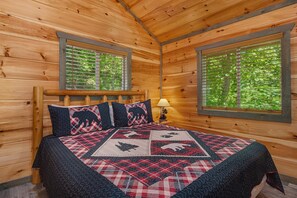 Une pièce de cabane en bois comprenant un lit, deux fenêtres et une table de chevet avec une lampe.