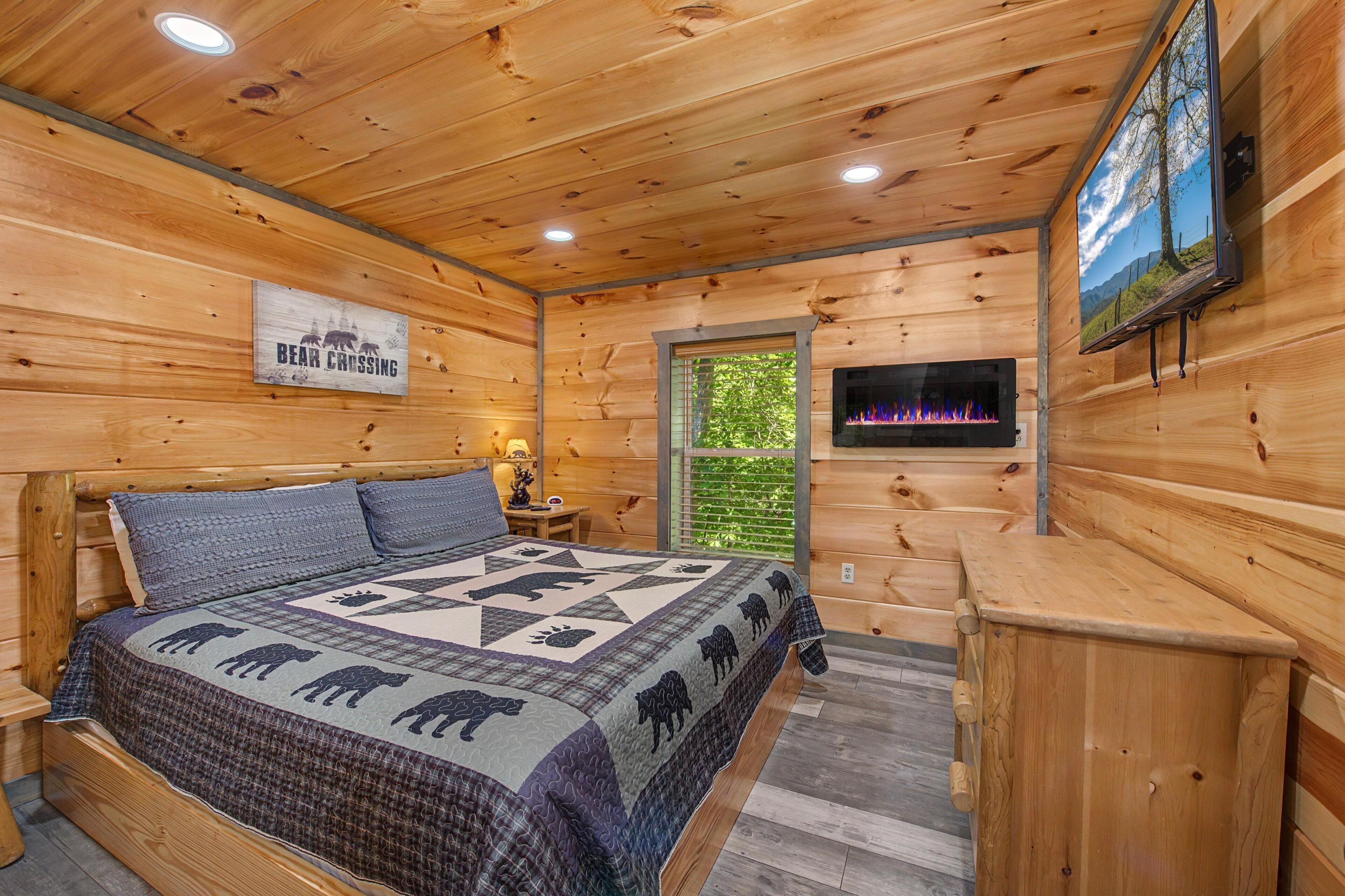A wooden cabin interior with a bed, a fireplace, a TV, and a window with a view of greenery.