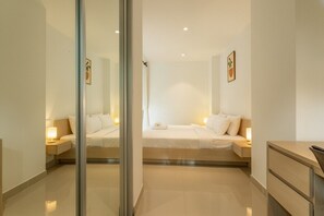 Ein modernes Hotelzimmer mit Glastrennwand, einem Bett mit weißer Bettwäsche, einem Nachttisch mit Lampe und einem kleinen Schreibtisch mit Schubladen.