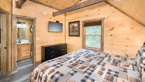 Una camera da letto con soffitto in legno, un letto con copriletto decorato, un comò, una televisione e una finestra vista alberi.