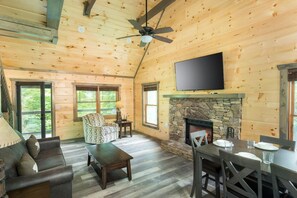 Ein gemĂŒtliches Blockhaus-Interieur mit Kamin aus Stein, HolzwĂ€nden, Deckenventilator und flachbildschirm-Fernseher ĂŒber dem Kamin.
