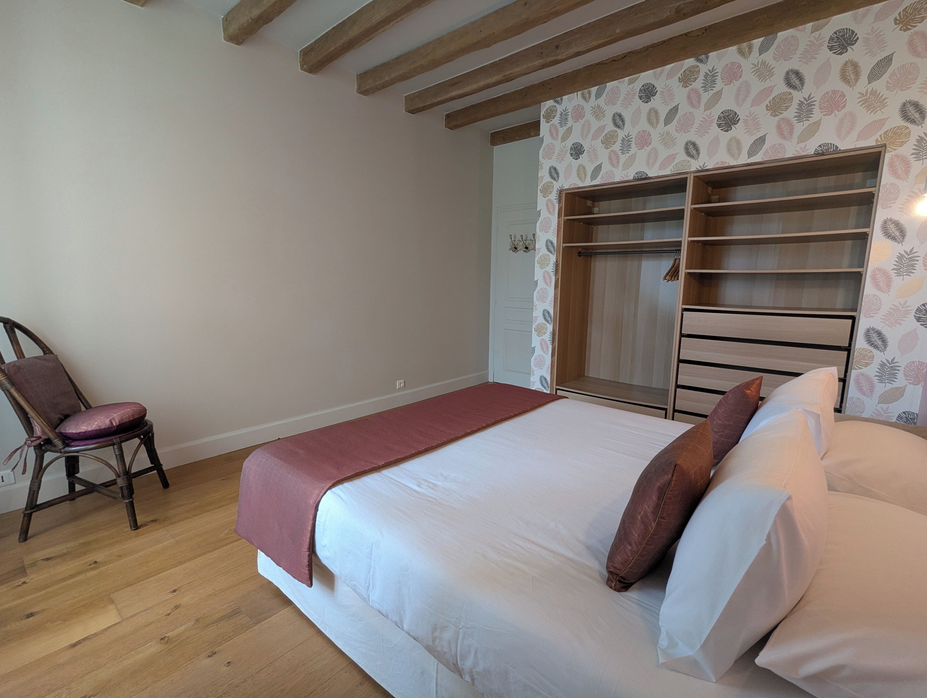Ein Schlafzimmer mit Holzboden, einem Bett mit weißer Bettwäsche, einem Holzstuhl und einem Kleiderschrank mit raurigem Wandpapier.