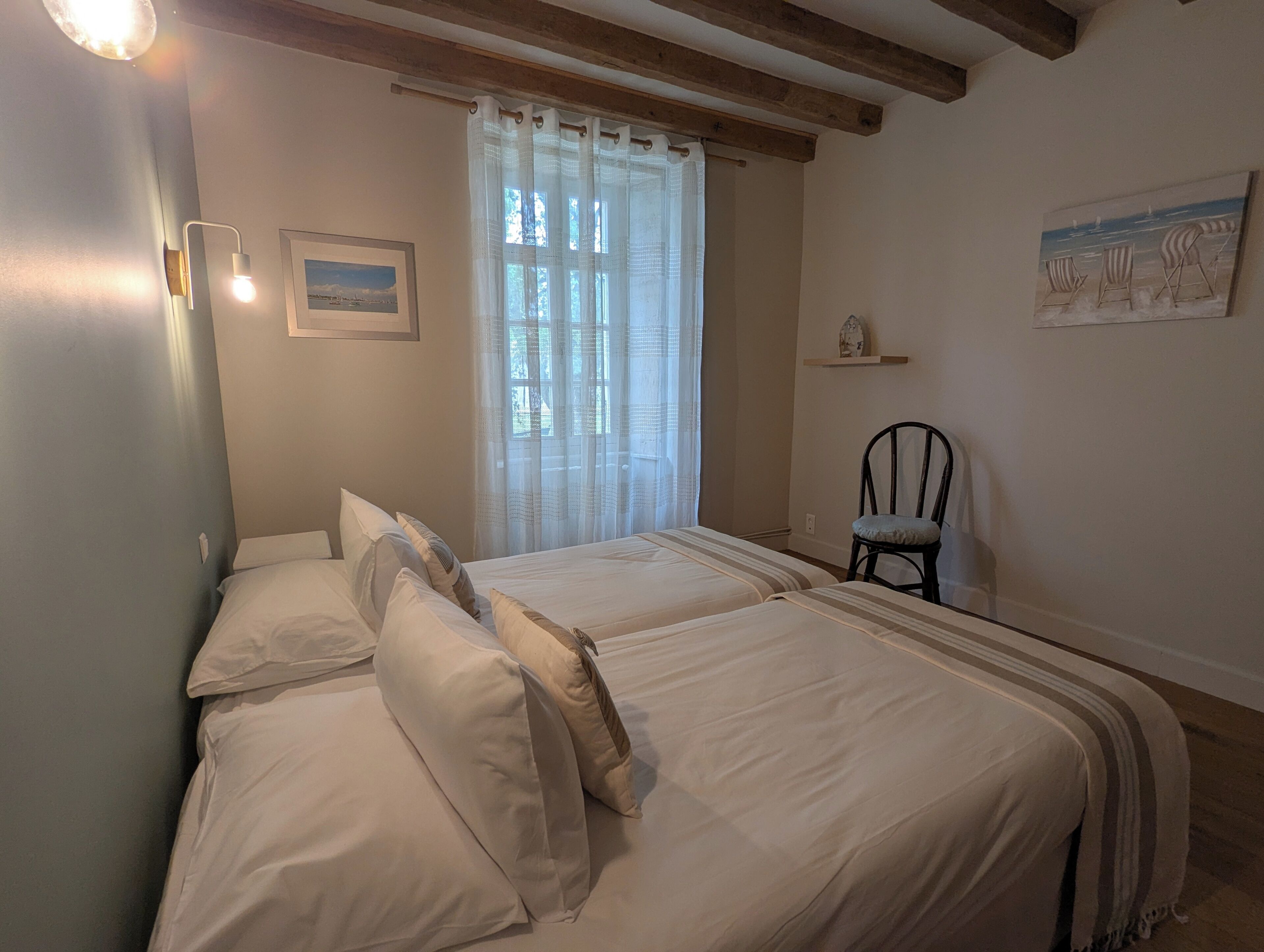 A room with two beds, a chair, a window with sheer curtains, a wall-mounted light, and a painting.