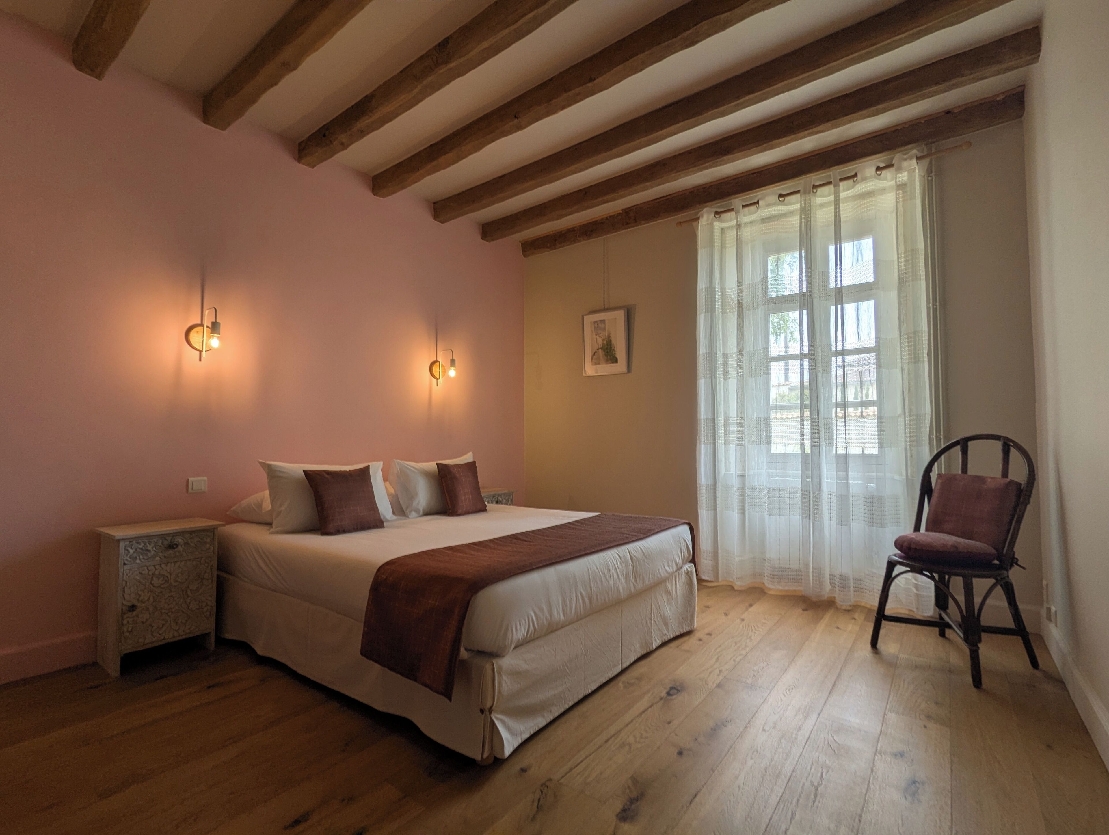 A bedroom with a wooden floor, a bed with pillows, a nightstand, a chair, and a window with curtains.