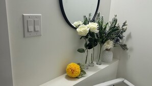 Un mur blanc orné d’un miroir rond, d’un interrupteur et d’une étagère avec des fleurs et un vase.