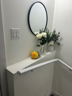 Una pared blanca con un espejo redondo, un interruptor de luz y una repisa con flores y un florero.