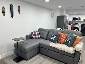 Una sala moderna con un sofá seccional gris, una mesita auxiliar y una cocina al fondo.