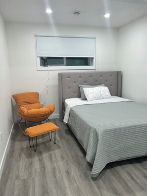 Un dormitorio con una cama, una silla, un banco, una ventana y una lámpara en el techo.