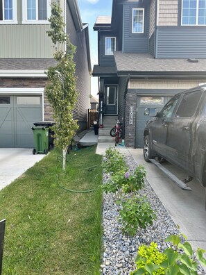 Eine Auffahrt zu einem Wohnhaus, mit einem geparkten SUV, einer grünen Mülltonne, einem kleinen Baum und einem Kiesweg.