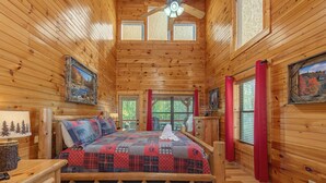 A log cabin bedroom with a wooden bed, a nightstand, a lamp, and a window with red curtains. - I Love View Too (Gatlinburg)