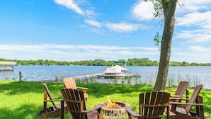 A lakeside setting with three Adirondack chairs facing a fire pit, a dock with a boat, and a tree. - Granite Lake Retreat w/ Private Dock (Annandale)